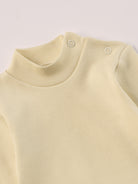 Dieser beige Stehkragenbody besteht aus hochwertiger, sorgfältig veredelter organischer Baumwolle. Die Stoffe sind sanft gefärbt und gebleicht unter Einhaltung strenger EU-Standards. Die nickelfreien Druckknöpfe an der Schulterpartie machen das An- und Ausziehen unkompliziert und schnell.

Innen sorgt die weich angeraute Oberfläche für angenehme Wärme, Feuchtigkeitsaufnahme und ein sanftes Hautgefühl. Der schützende Stehkragen hält den empfindlichen Halsbereich warm und reduziert Zugluft.